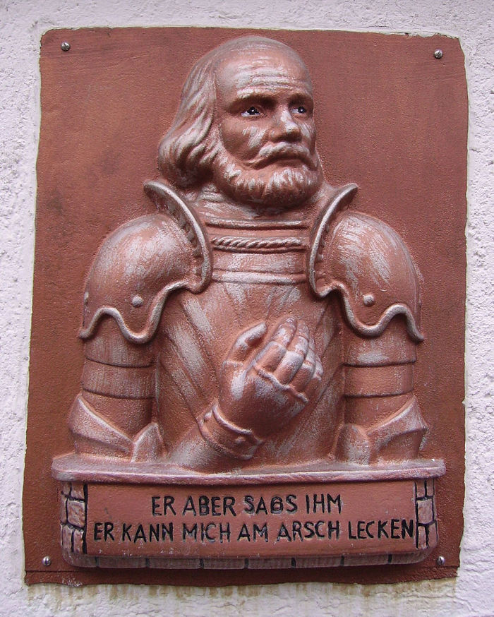 Reliefskulptur eines Ritters, darunter in Großbuchstaben der Spruch »Er aber sags ihm er kann mich am Arsch lecken«.