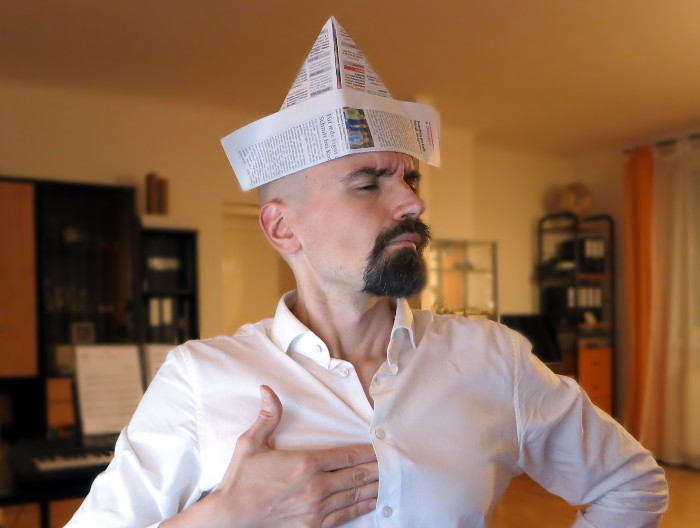 Ich mit einem Origami-Hut aus Zeitungspapier. Ich mache einen ernsten Gesichtsausdruck mit gerunzelter Stirn und schiebe mir vier Finger unter die Knopfleiste meines Hemdes (– in dieser Pose werden oft Verrückte dargestellt, die Napoleon imitieren).