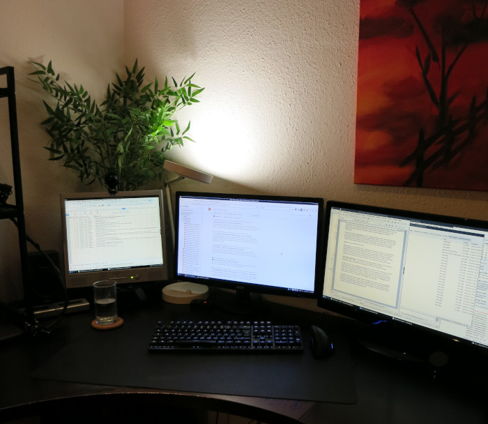 Schreibtisch mit drei Monitoren vor einer weißen Wand. Hinter den Monitoren steht eine Schreibtischlampe, die nach oben gegen die Wand gerichtet ist.