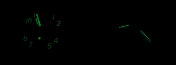 Die nachleuchtenden Elemente von zwei Uhren im Dunkeln. Links sieht man deutlich zwei Zeiger und acht Zahlen am Zifferblatt, rechts sieht man nur zwei dünne Zeiger.