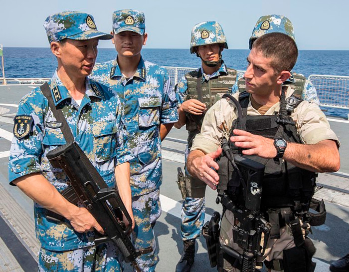 Soldaten auf einem Schiff vor dem Ozean. Die meisten tragen eine Uniform mit einem vorwiegend blauen Muster.