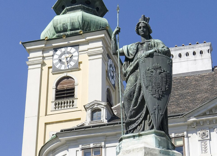 Frauenstatue mit Speer und Schild vor einem Uhrturm.