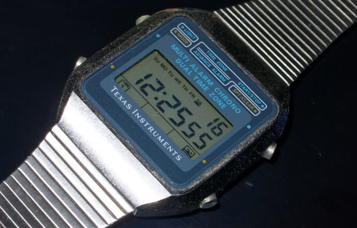 Digitaluhr mit schwarzen Siebensegment-Ziffern auf einem gräulichen Hintergrund.