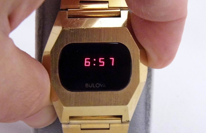 Armbanduhr, die rot leuchtende Digitalziffern anzeigt, während sie zwischen zwei Fingern gedrückt wird.