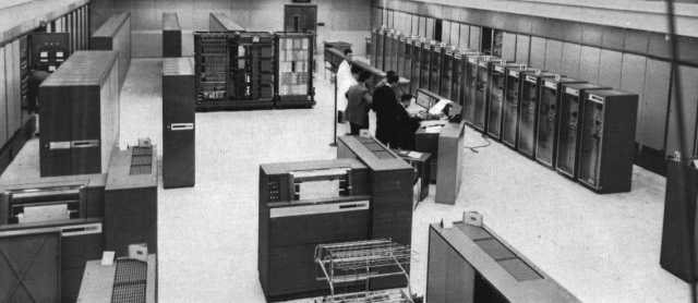 Schwarzweiß-Foto von Großrechner, der vorwiegend aus einem großen Raum voll hoher Computerschränke besteht.