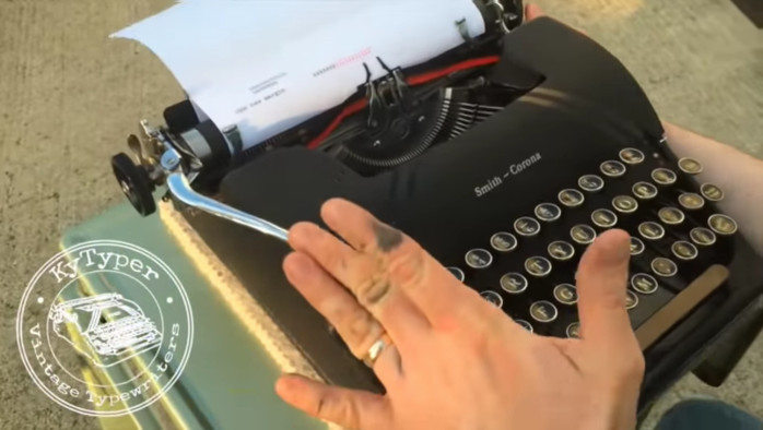 Eine Hand mit schwarzem Fleck am Zeigefinger schiebt am Hebel einer mechanischen Schreibmaschine den Wagen zurück.