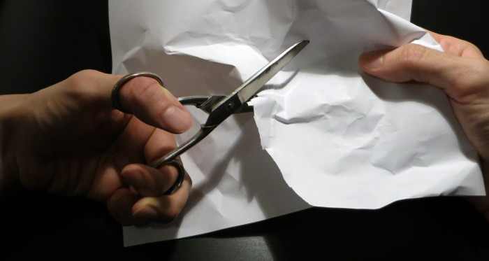 Zerknittertes und schief geschnittenes Blatt Papier unter einer linkshändig gehaltenen Schere.