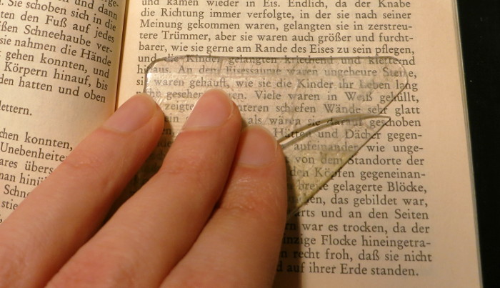 Drei Finger halten ein flaches, transparentes, abgestumpftes und zerbrochenes Ding unter einer Textzeile in einem Buch.